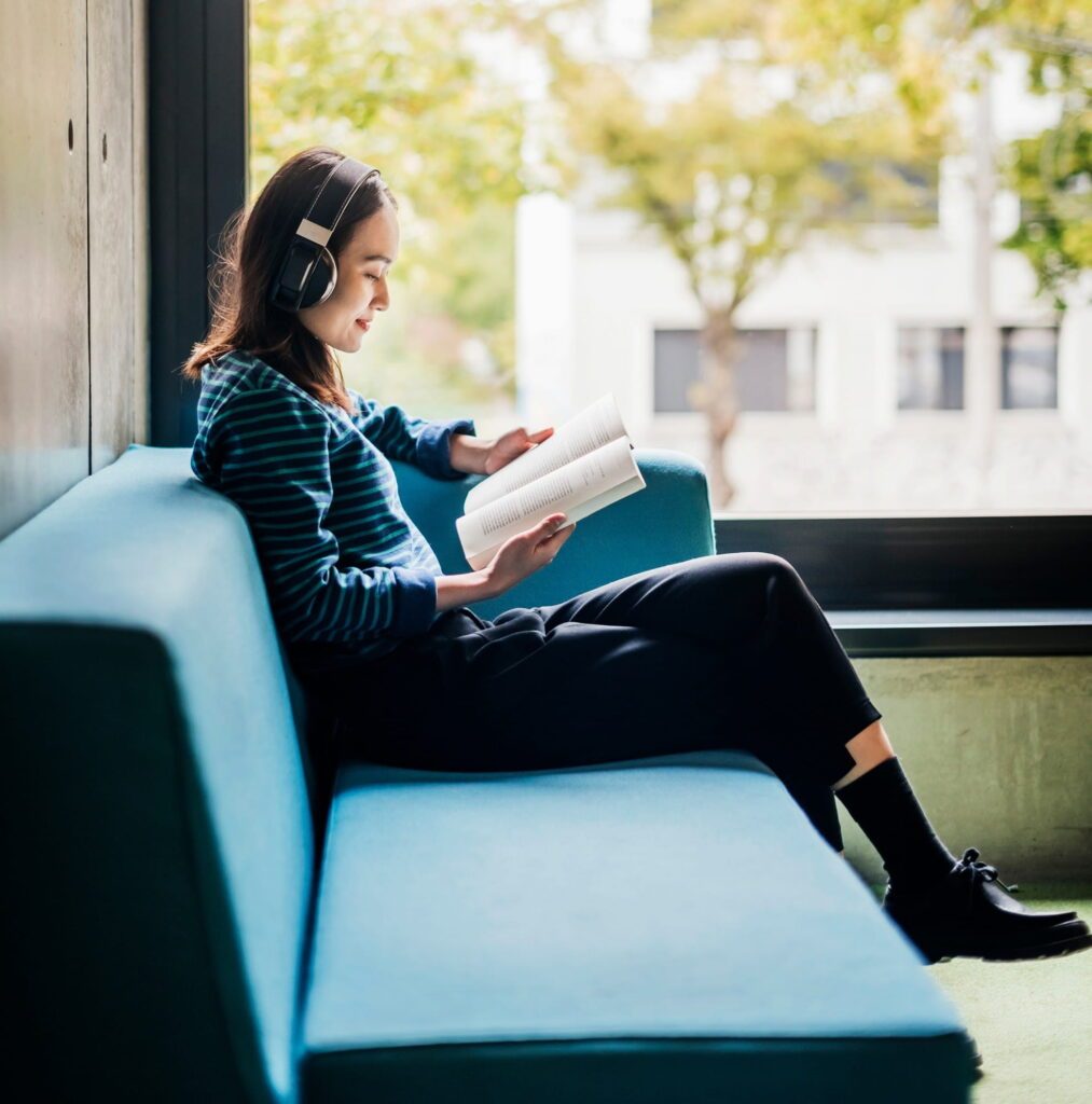 A person sits on a blue couch, reading a book while wearing headphones, with greenery visible outside a window.