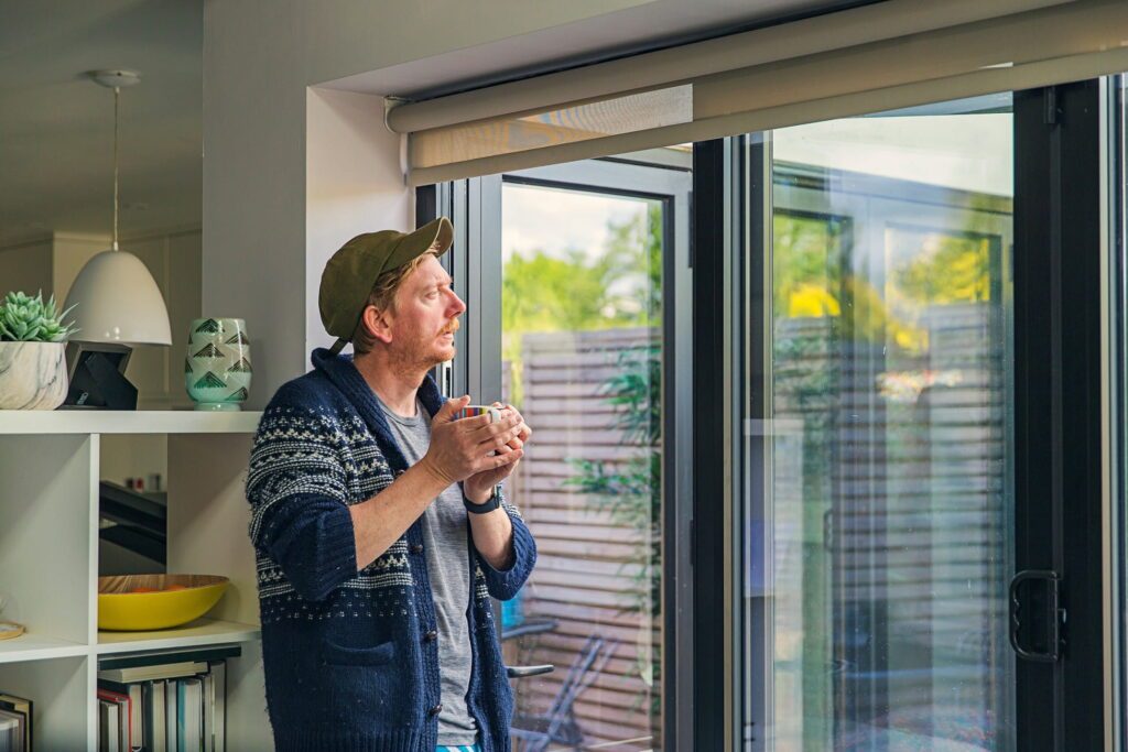 A person in a cozy sweater stands by a large window, holding a colorful mug and admiring the view outside.